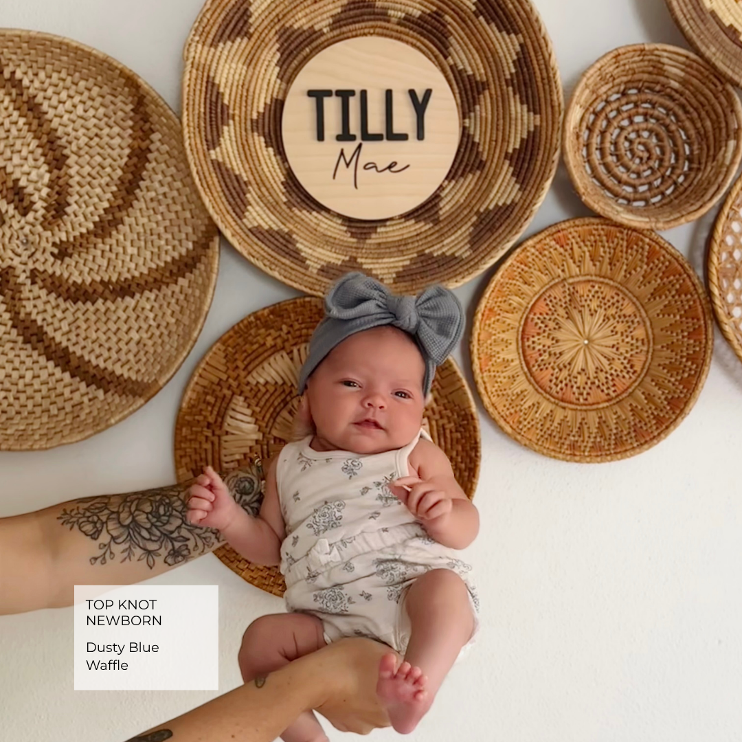 A baby wearing a dusty blue top knot headband and a light patterned romper lies on a woven mat, surrounded by other natural woven baskets and plates on a white background. A text box in the lower left reads 'TOP KNOT NEWBORN Dusty Blue Waffle'.