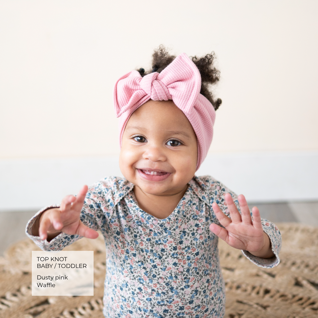 A smiling toddler wearing a dusty pink ribbed top knot headband with a large bow, paired with a blue and pink floral long-sleeve romper. The toddler is posing with hands out on a neutral background with a text overlay in the bottom left corner reading 'TOP KNOT BABY / TODDLER Dusty pink Waffle'.