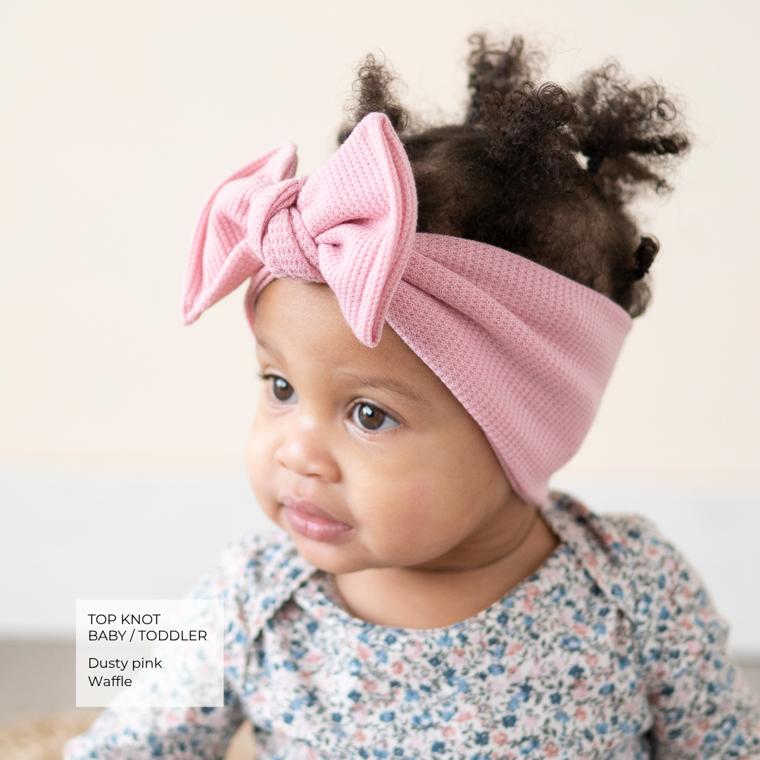 A baby wearing a dusty pink knitted top knot headband with a large bow, paired with a floral long-sleeve top. A text overlay in the bottom left reads 'TOP KNOT BABY / TODDLER Dusty pink Waffle'.