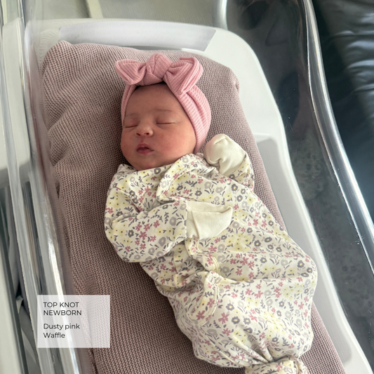 A sleeping newborn baby wearing a dusty pink top knot headband and a matching floral footed swaddle blanket, lying in a white bassinet. A text overlay on the bottom left reads 'TOP KNOT NEWBORNS Dusty pink Waffle'.