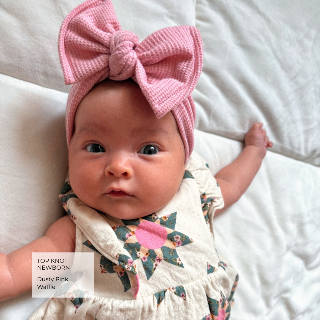 A newborn baby lying on white bedding, wearing a dusty pink knitted top knot headband with a large bow and a cream floral bib. A text overlay in the bottom left corner reads 'TOP KNOT NEWBORNS Dusty Pink Waffle'.