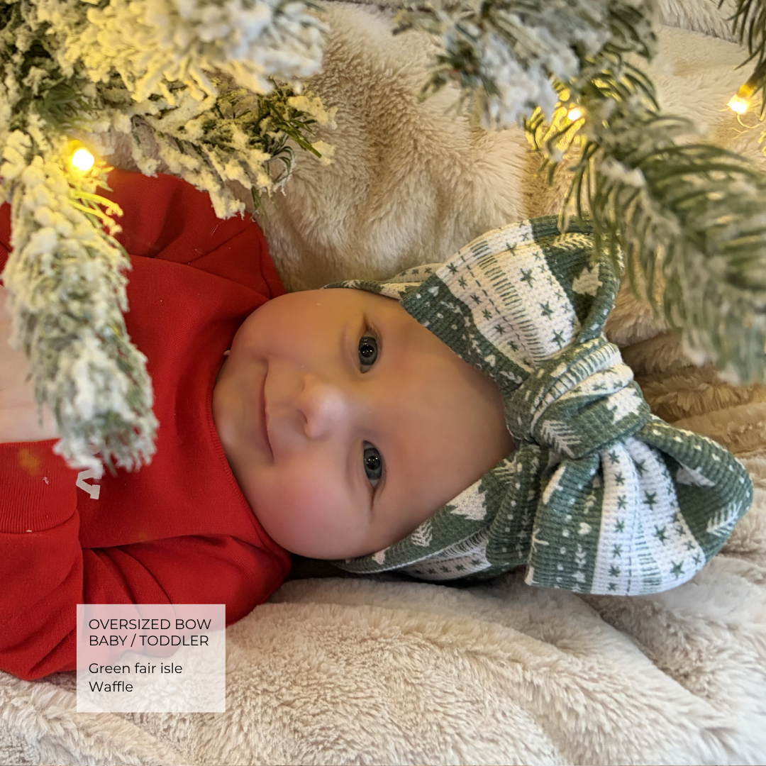 A baby wearing an oversized green and white patterned bow headband lies on a soft cream blanket under a decorated Christmas tree with warm lights. A text label in the lower left reads 'Oversized Bow Baby / Toddler Green fair Isle Waffle'.