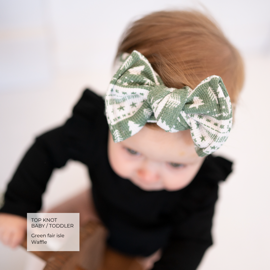 A toddler wearing a green and white patterned top and a matching green fair isle waffle top knot baby headband, photographed against a bright white background.