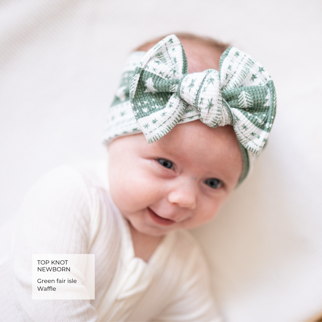 A smiling newborn baby wearing a green and white patterned knit top knot bow headband, lying on a soft white blanket. A text overlay in the bottom left reads 'TOP KNOT NEWBORN Green fair isle Waffle'.