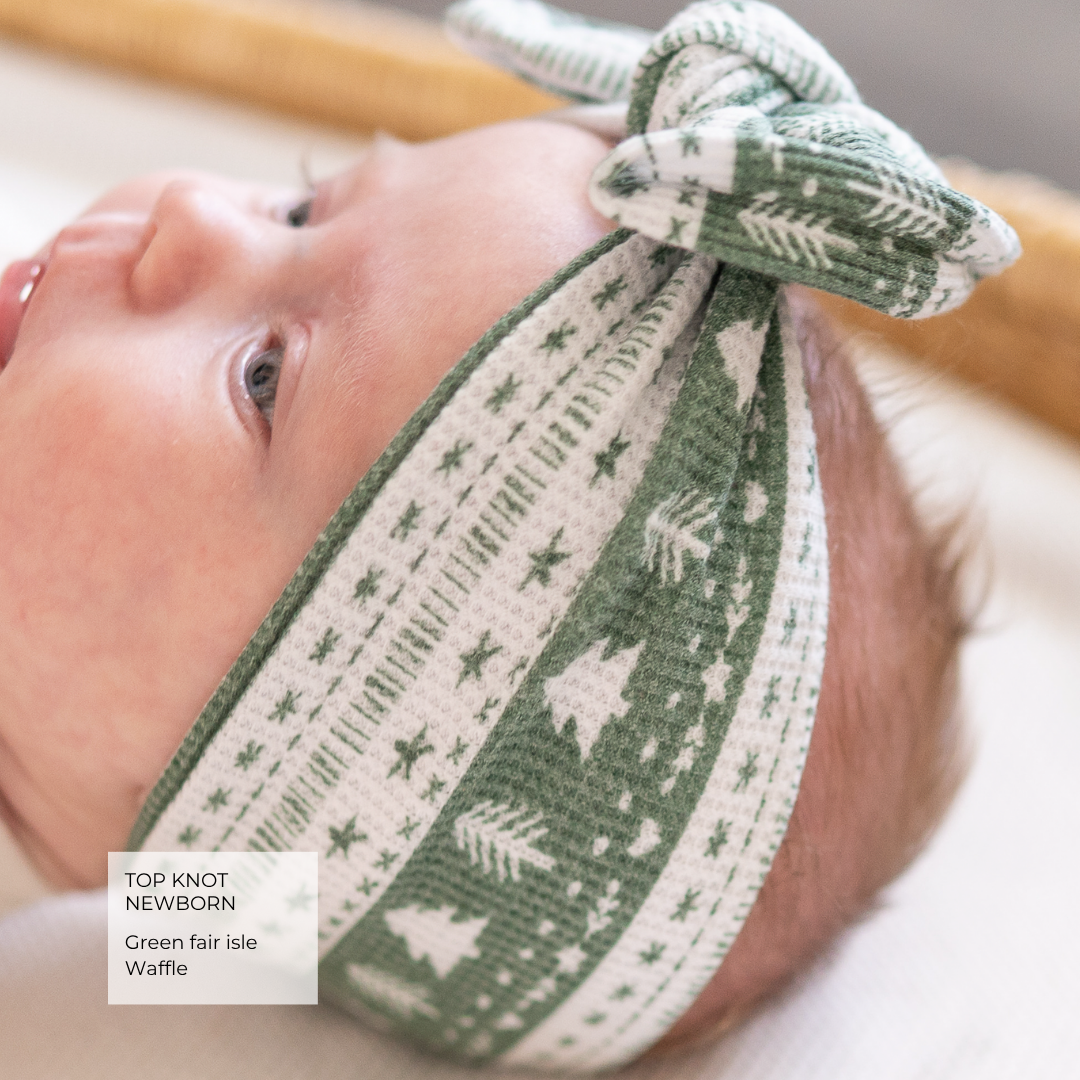 A close-up of a newborn baby wearing a green and white patterned knit top knot headband with a bow, lying in a crib.