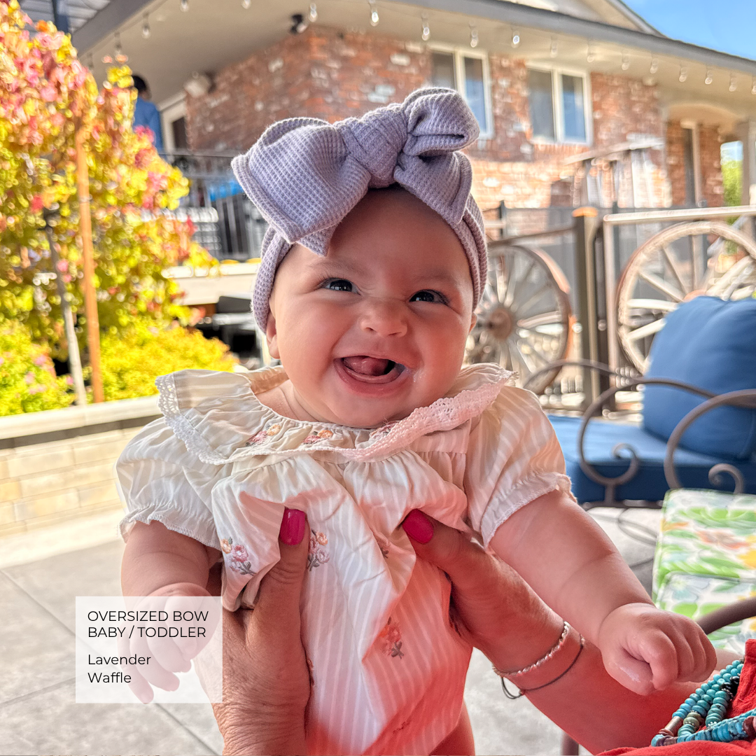 A smiling baby wearing an oversized lavender waffle-textured bow headband and a light pink ruffled dress, held up by an adult outdoors in front of a brick house.