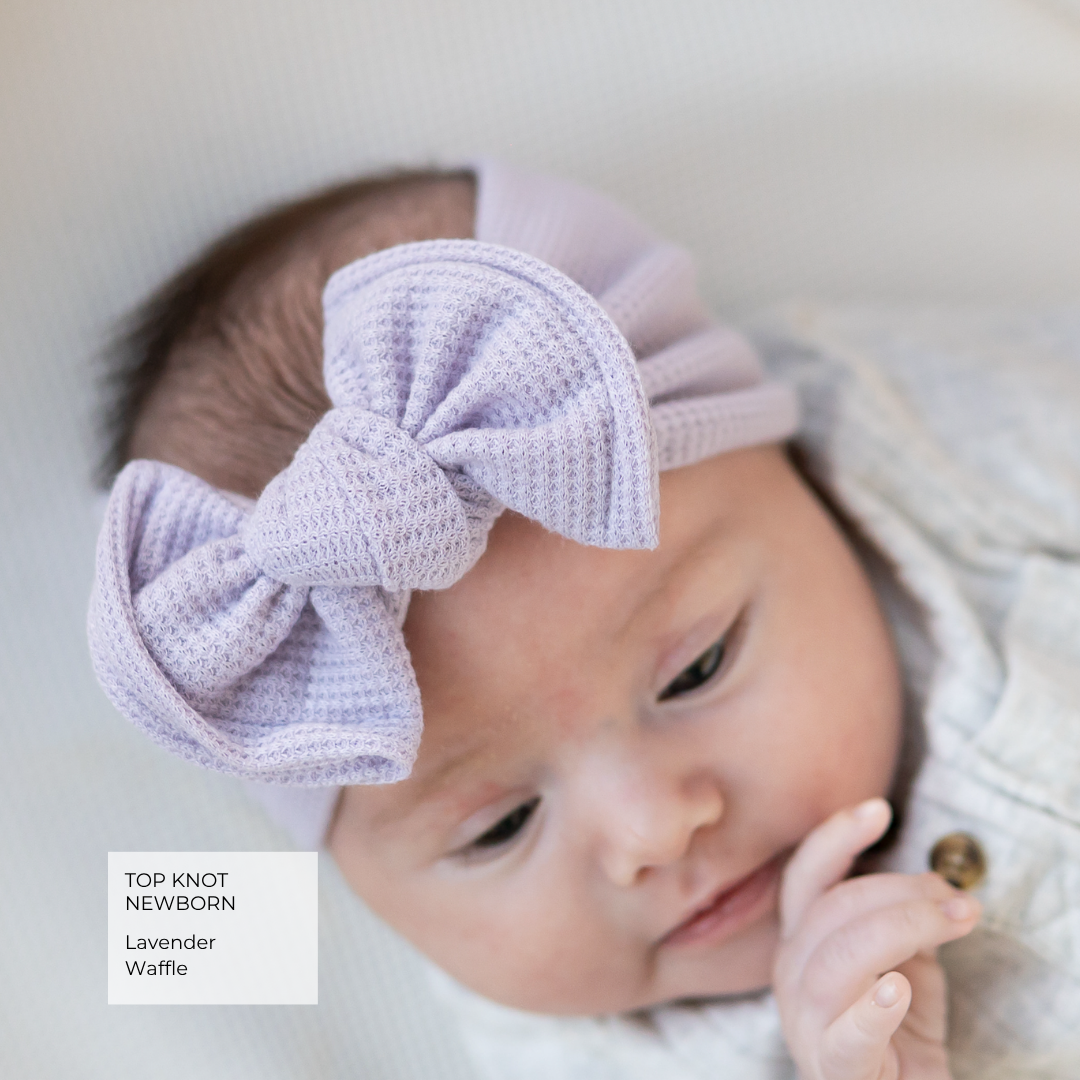 A close-up of a newborn baby wearing a lavender waffle-knit top knot headband with a large bow. The baby is dressed in a light knit outfit, lying on a soft, light-colored surface.