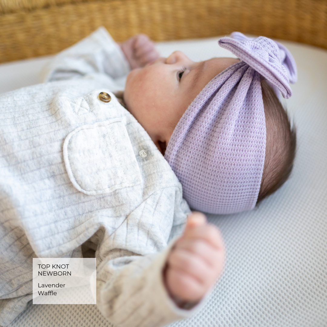 A newborn baby lying on a textured white surface, wearing a lavender waffle knit top knot headband with a bow and a light gray quilted romper. A text overlay in the bottom left corner reads 'TOP KNOT NEWBORN Lavender Waffle'.