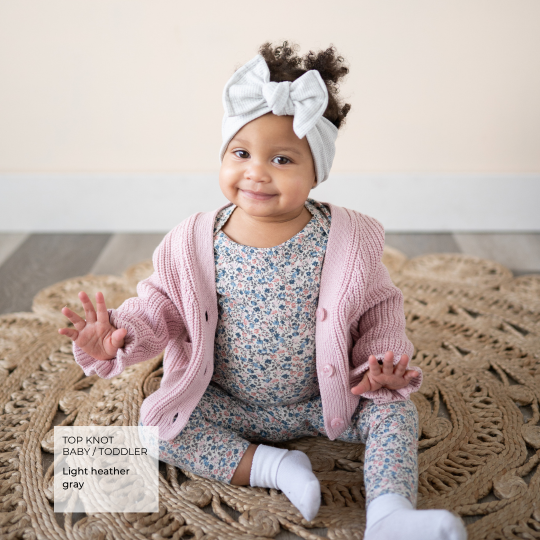 baby wearing a pink cardigan and gray bow, sitting on a boho rug.