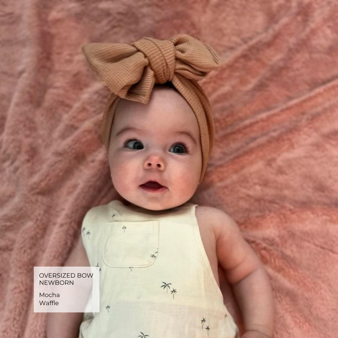 A newborn baby lying on a textured dusty pink blanket, wearing an oversized waffle-knit bow headband in mocha brown and a cream sleeveless romper with tiny black palm tree prints. A text overlay in the bottom left corner reads 'Oversized Bow Newborn Mocha Waffle'.