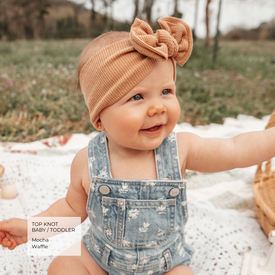 A baby wearing a waffle-knit top knot headband in mocha brown and denim overalls, sitting outdoors on a white blanket. A text overlay in the bottom left reads 'TOP KNOT BABY / TODDLER Mocha Waffle'.