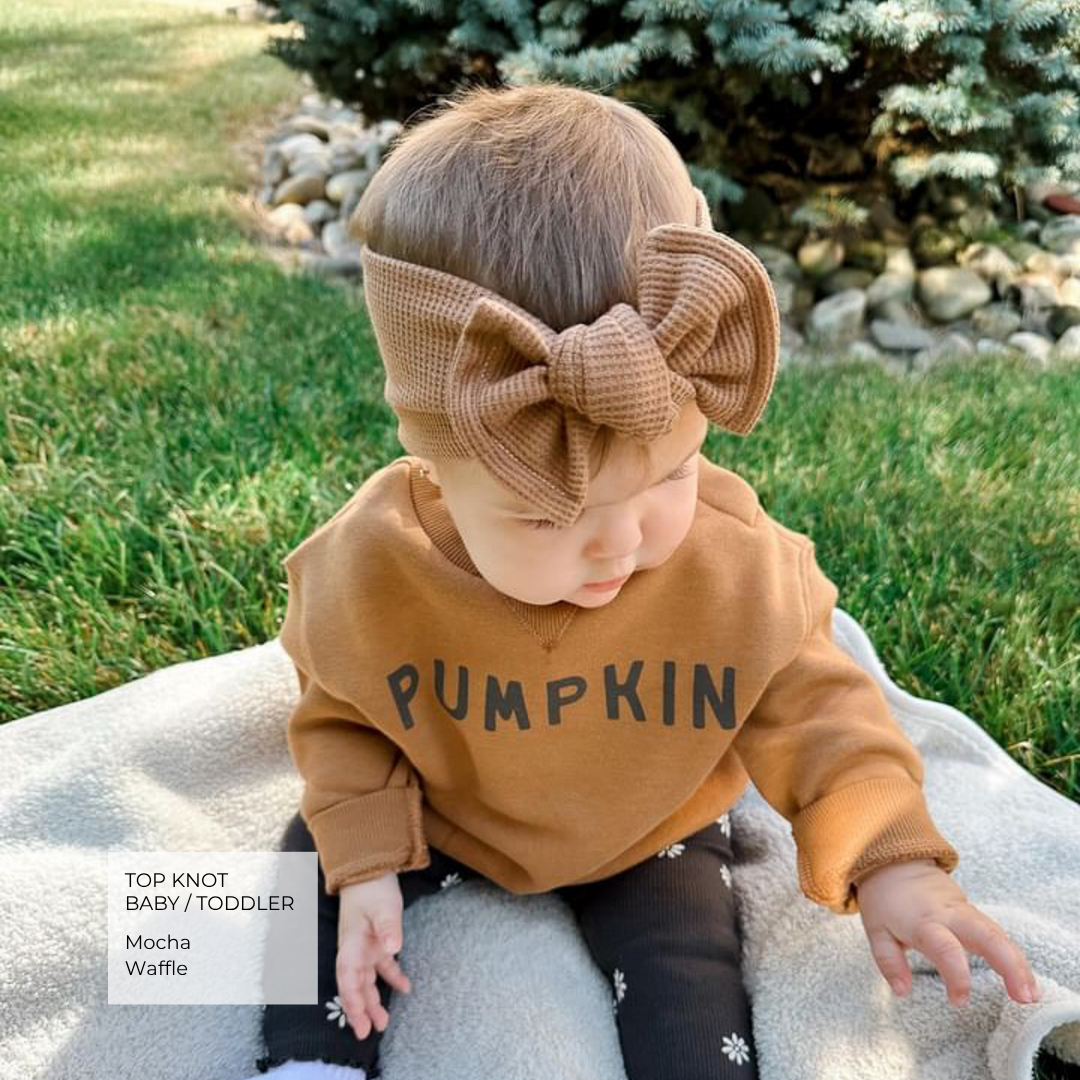 baby wearing a mocha waffle top knot bow and brown sweatshirt that says PUMPKIN.