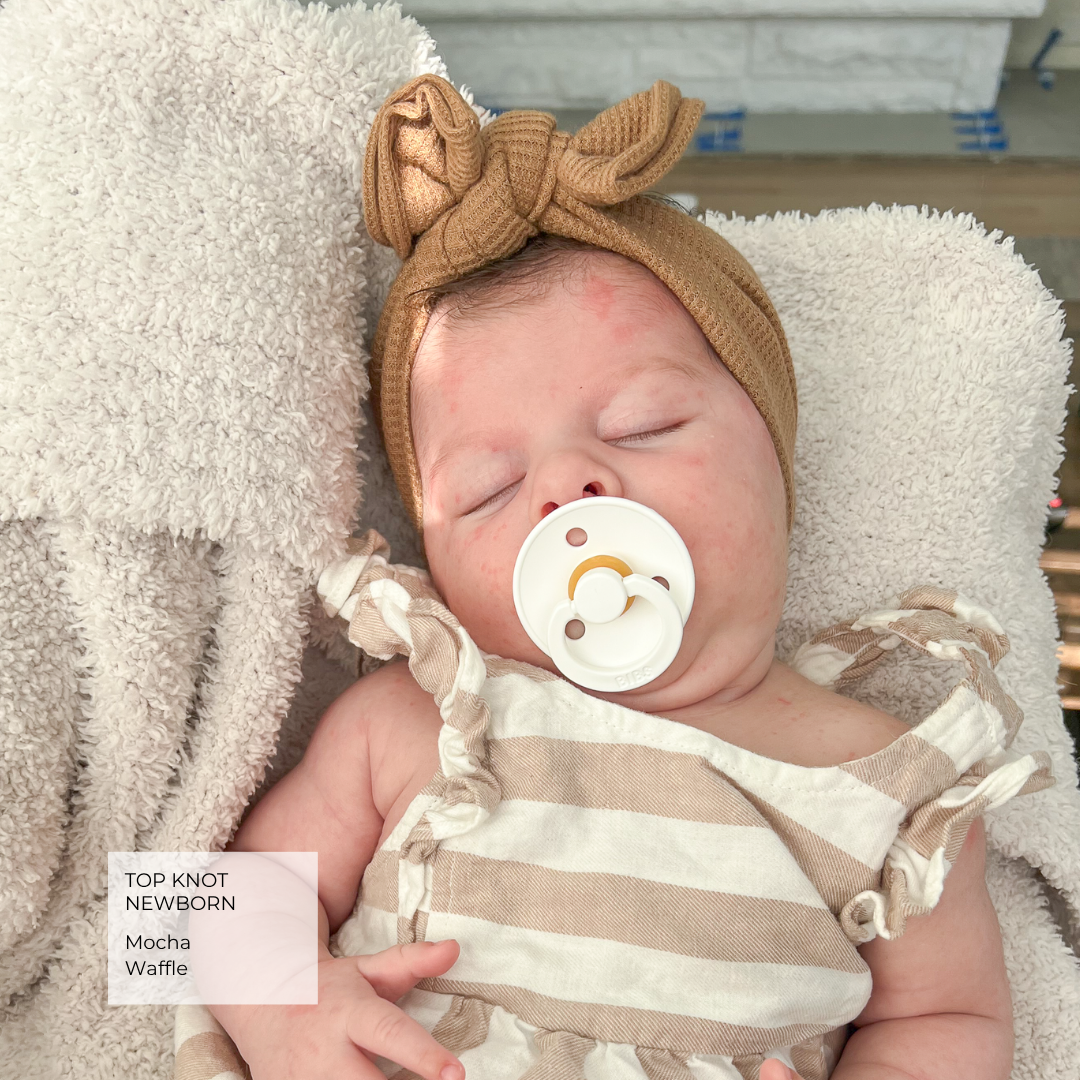 A sleeping newborn baby wearing a tan knit top knot headband with a bow, a beige and white striped romper, and a white pacifier, resting on a fluffy cream blanket. A text overlay in the bottom left reads 'TOP KNOT NEWBORN Mocha Waffle'.