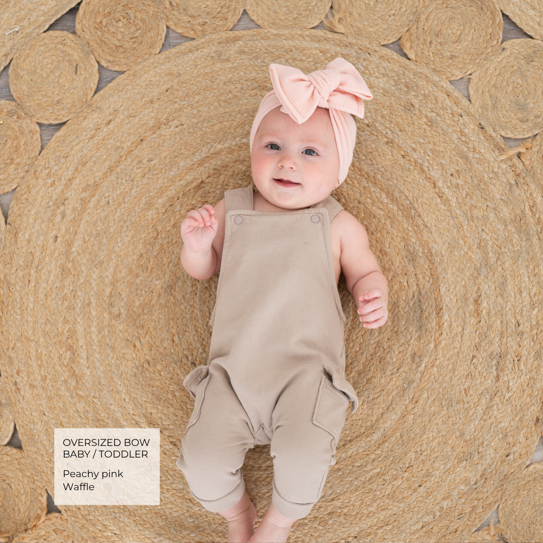 A baby lying on a round woven jute rug, wearing a peach pink oversized bow headband and a light beige sleeveless romper. A text box in the bottom left corner reads 'OVERSIZED BOW BABY / TODDLER Peachy pink Waffle'.