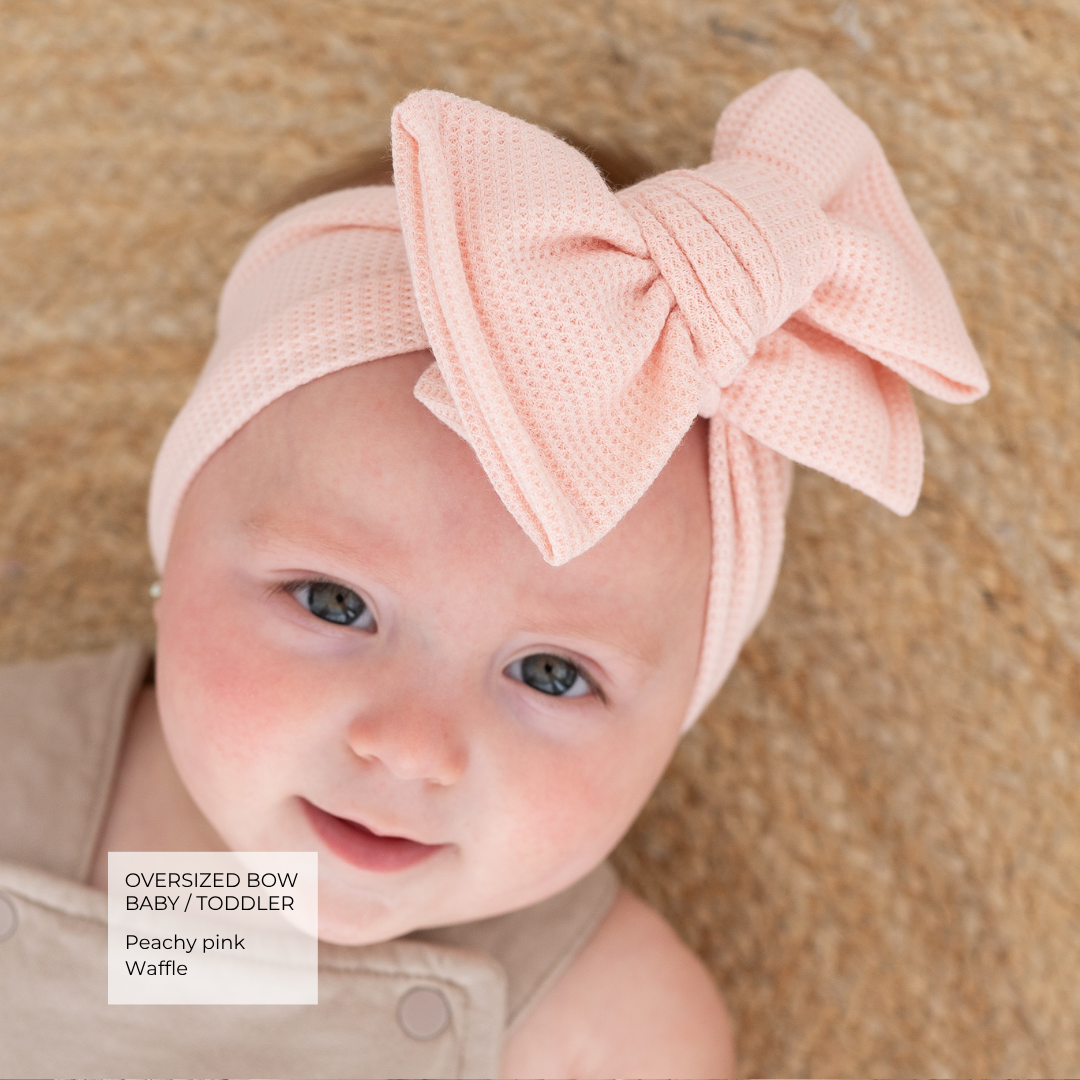 A baby wearing a peachy pink oversized bow waffle headband, lying on a textured beige blanket. A text overlay in the bottom left corner reads 'Oversized Bow Baby / Toddler Peachy pink Waffle'.