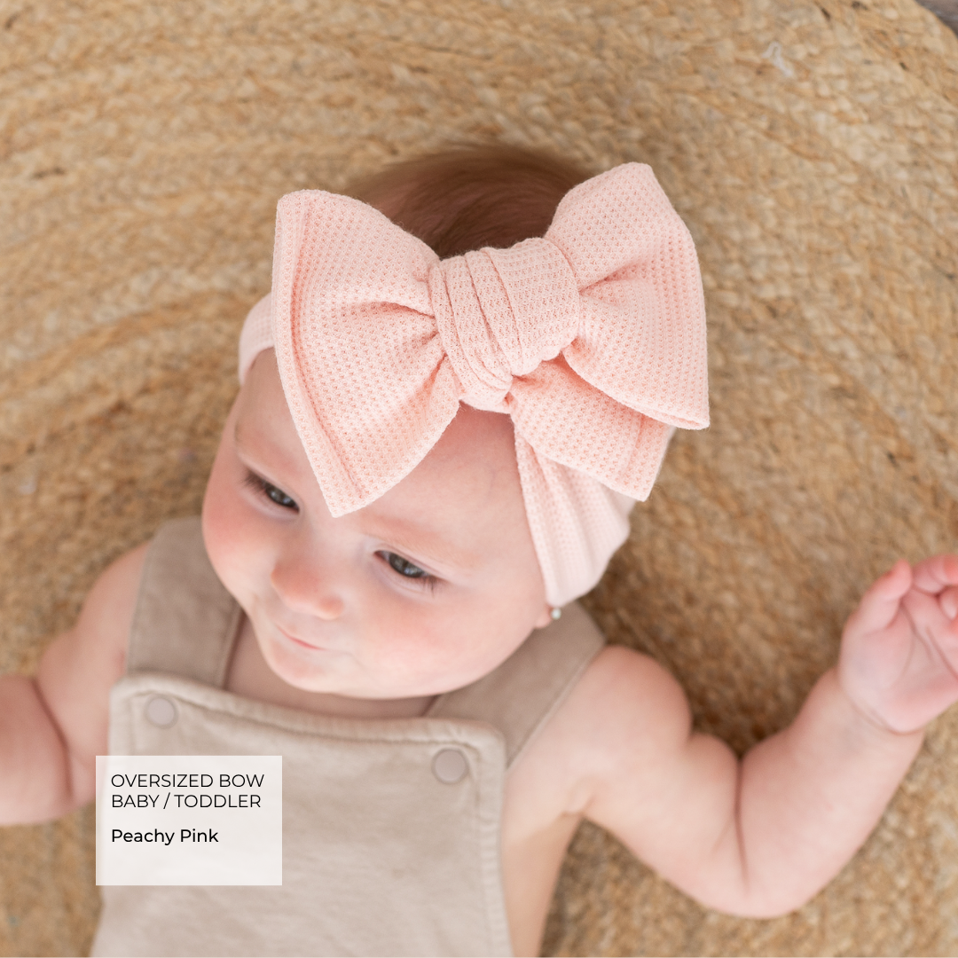 Baby wearing a peach-colored oversized bow headband on a textured brown background