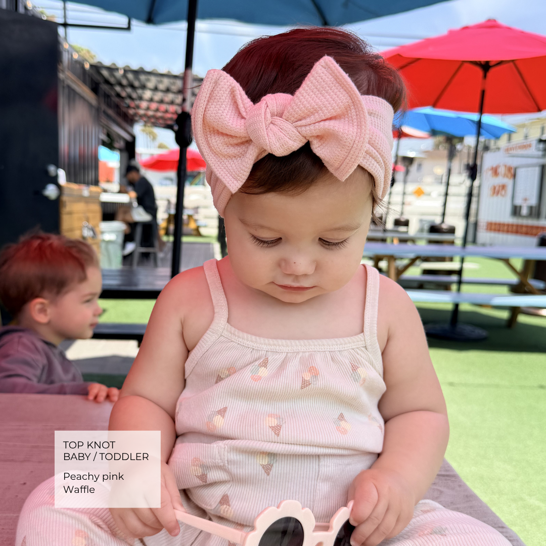 A toddler wearing a large peachy pink top-knot bow headband and a matching light pink ice cream-patterned ribbed tank top, sitting outdoors under a patio umbrella.
