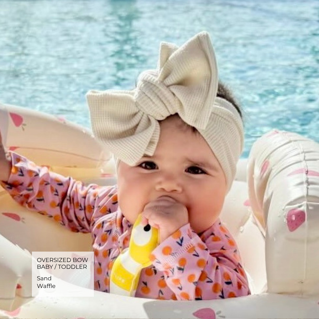 A baby wearing an oversized cream-colored ribbed bow headband and a pink citrus-patterned long-sleeve shirt, sitting in a white inflatable pool float with blue water in the background.