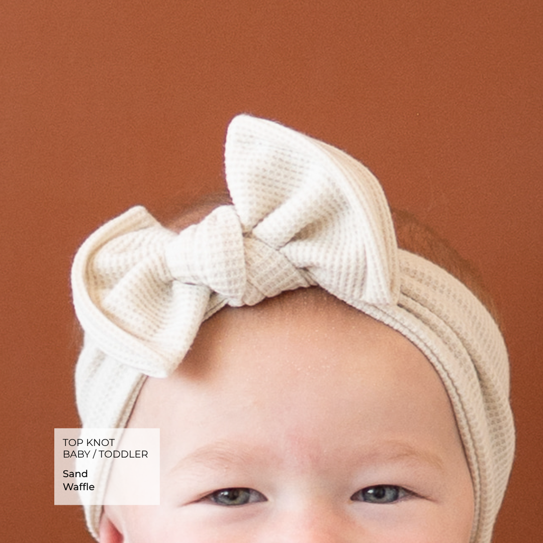 A close-up of a baby wearing a textured cream waffle-knit top knot headband with a large bow, set against a solid terracotta brown background. A text overlay in the bottom left reads 'TOP KNOT BABY / TODDLER Sand Waffle'.