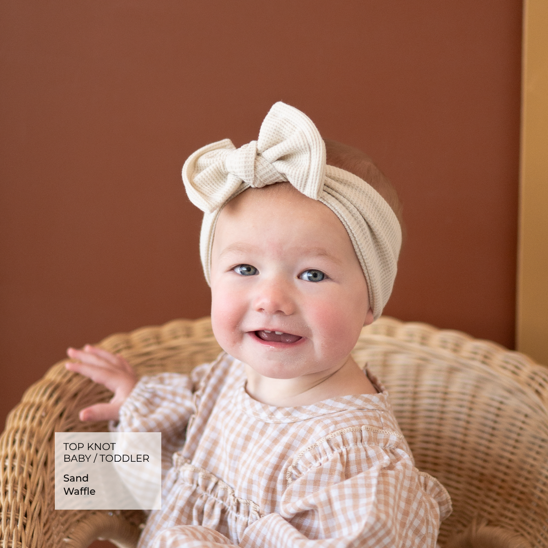 A smiling baby toddler wearing a sand-colored top-knot headband and a beige-and-white gingham dress, sitting in a wicker chair against a warm brown background.