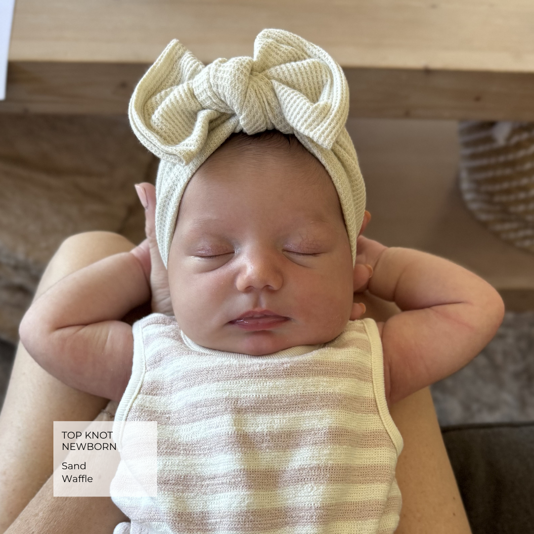 A sleeping newborn baby wearing a cream-colored knitted top knot headband with a large bow and a matching striped swaddle, held on an adult's lap. A text overlay in the bottom left reads 'TOP KNOT NEWBORNSand Waffle'.