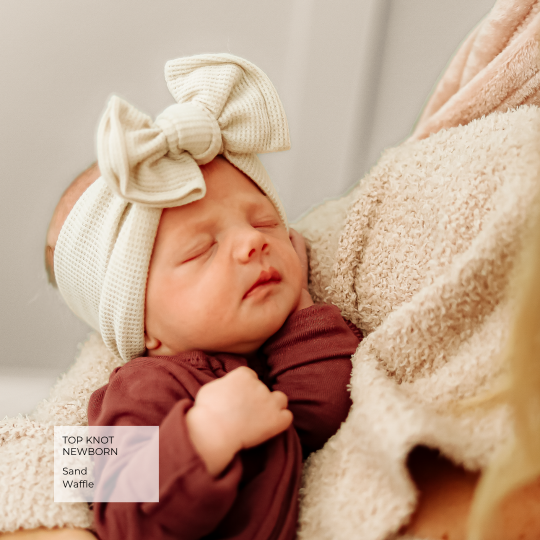 A sleeping newborn baby wearing a cream waffle-knit top knot headband with a large bow and a burgundy long-sleeve outfit, nestled in a beige blanket. A text overlay in the bottom left reads 'TOP KNOT NEWBORNS Sand Waffle'.