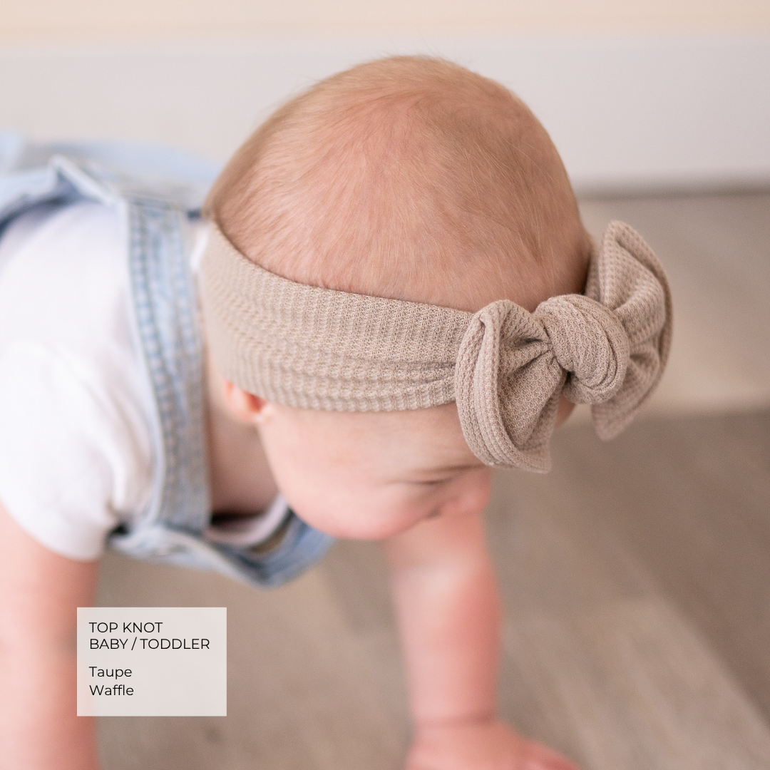 A baby wearing a taupe knit top-knot headband, shown from behind as they crawl on a light wooden floor. A text overlay in the bottom left reads 'TOP KNOT BABY / TODDLER Taupe Waffle'.