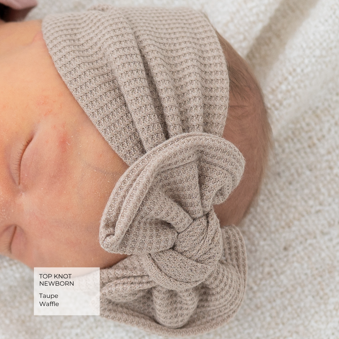 A newborn baby wearing a taupe waffle-knit top knot headband with a large bow, lying on a textured white blanket. A text label in the bottom left reads 'TOP KNOT NEWBORN Taupe Waffle'.