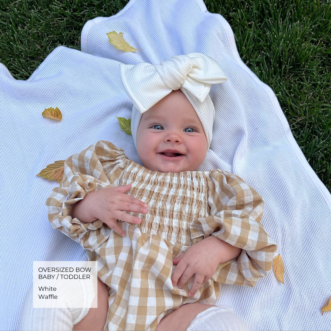 Baby wearing a white waffle oversized bow and a brown gingham onesie. lying in the grass on a white blanket with autumn leaves.