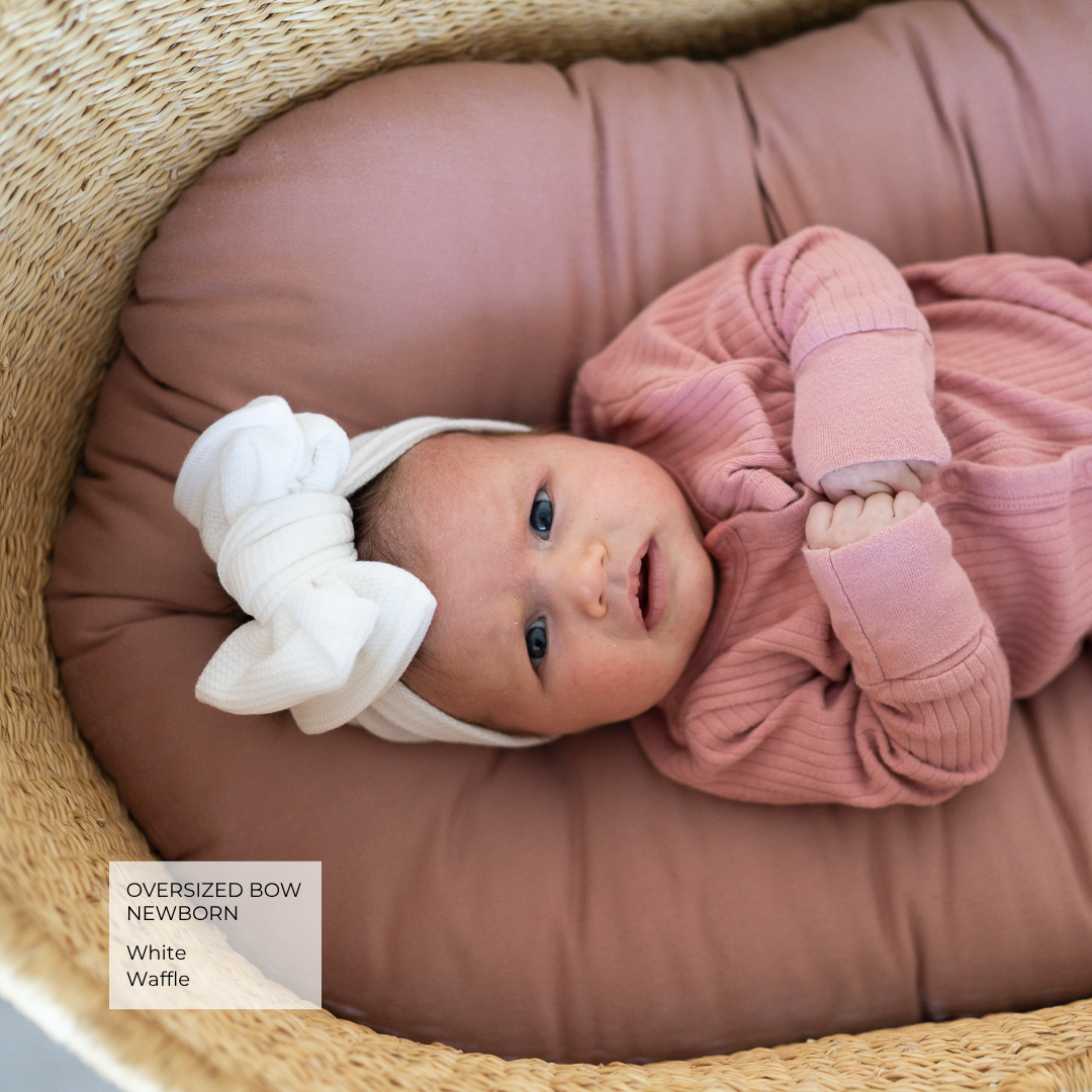 A baby wearing an oversized white bow headband and a dusty pink ribbed long-sleeve bodysuit, lying on a mauve cushion inside a woven basket. A text label in the lower left reads 'OVERSIZED BOW NEWBORN White Waffle'.