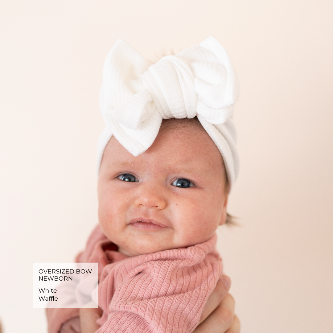 A newborn baby wearing an oversized white fabric bow headband and a pink ribbed wrap, held by an adult against a soft beige background. A text overlay in the bottom left reads 'OVERSIZED BOW NEWBORN White Waffle'.