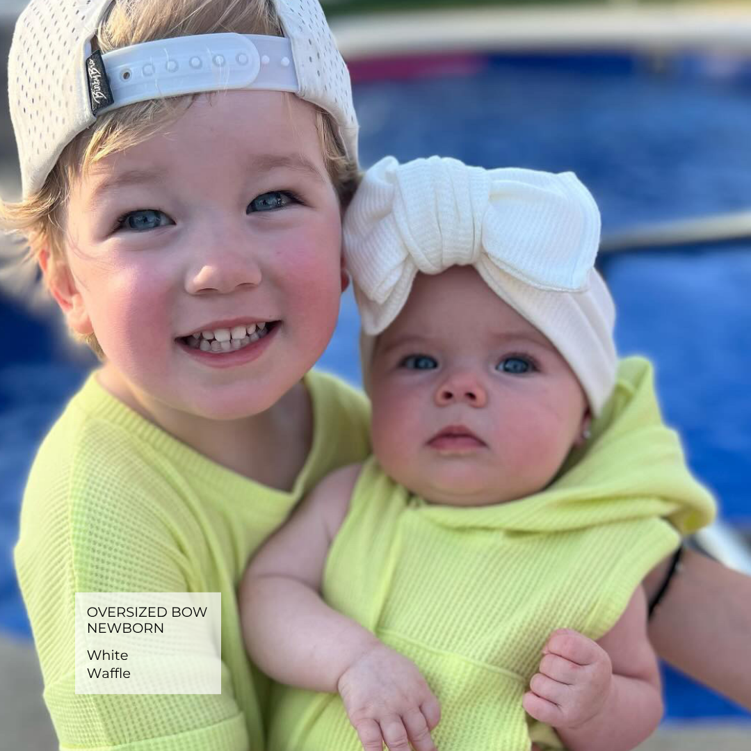 Two toddlers wearing matching pale yellow waffle-knit outfits and white waffle-knit accessories, including an oversized bow headband and a matching cap, posed outdoors in front of a blue swimming pool.