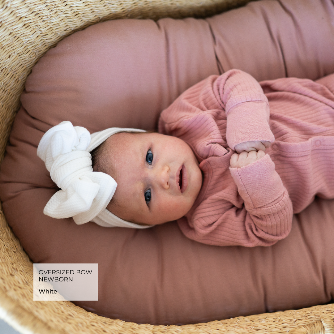 baby in Moses basket wearing a pink outfit and white oversized bow.