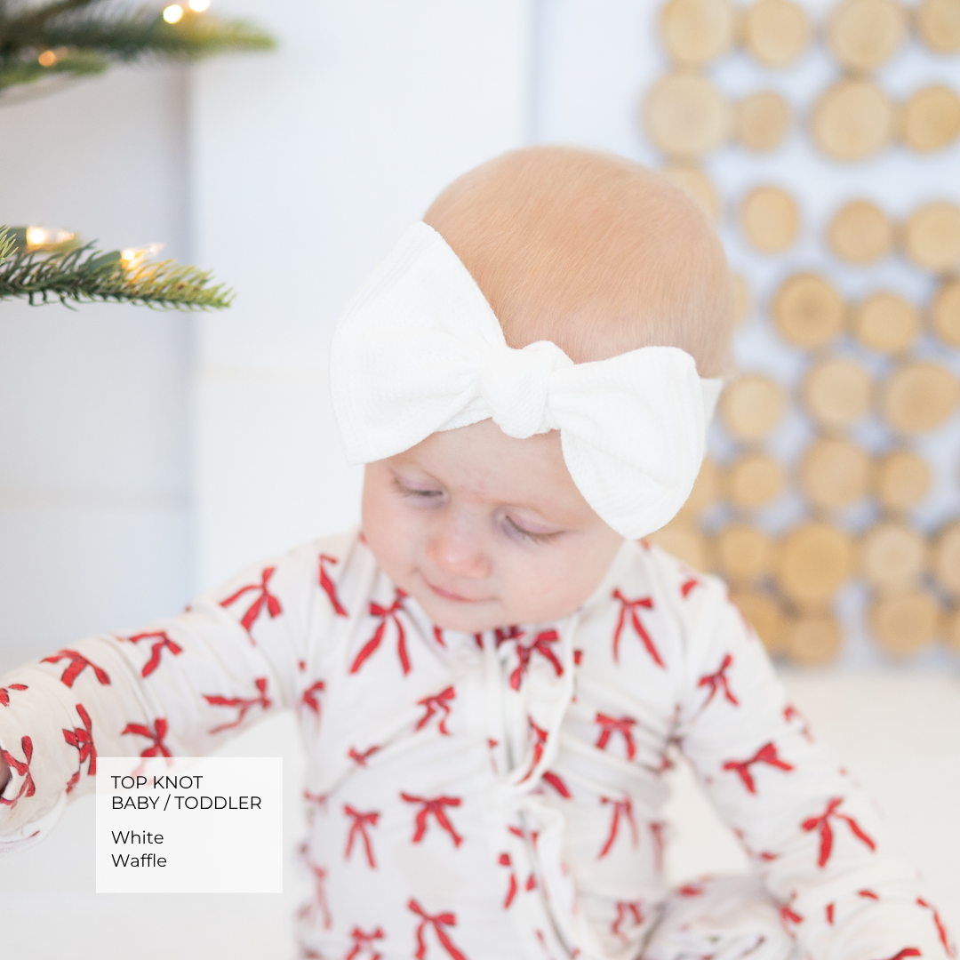 Baby wearing red bow Christmas pajamas and a white waffle top knot bow.