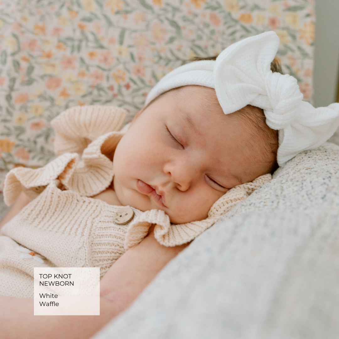 A sleeping newborn baby wearing a white knit top and matching white bow headband, resting on a textured blanket with a floral-patterned background. A text label in the corner reads 'TOP KNOT NEWBORN White Waffle'.