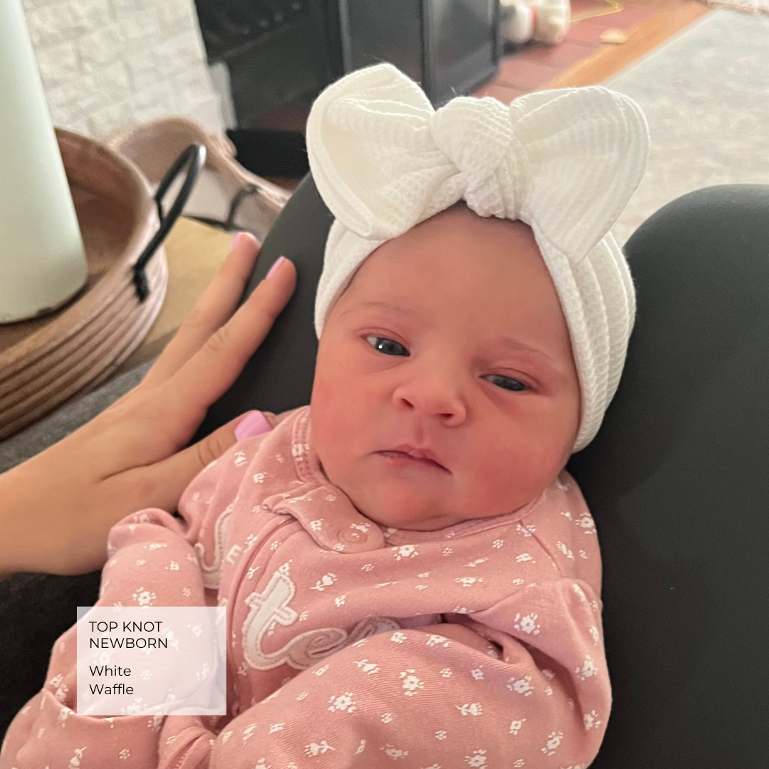 A newborn baby wearing a white ribbed top knot headband with a large bow and a pink patterned onesie, held by an adult in a home setting. A text overlay in the bottom left reads 'TOP KNOT NEWBORNS White Waffle'.