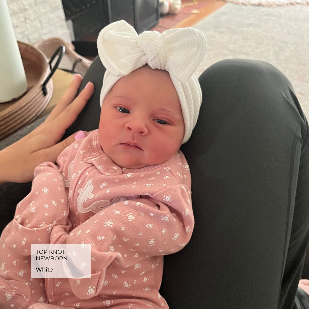 Newborn baby wearing a white headband with a bow, sitting on a person's lap.