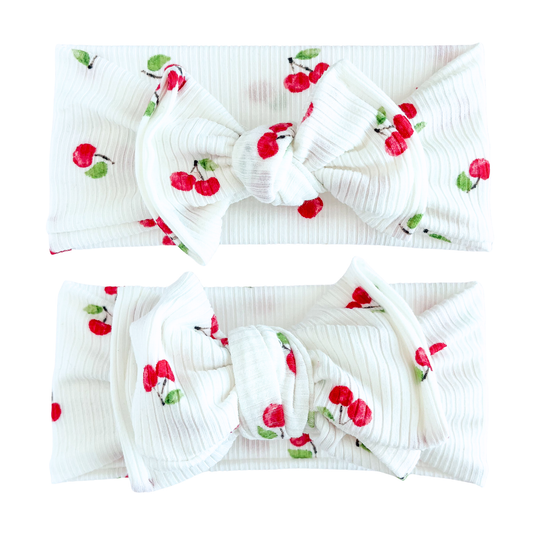 flat lay of two white ribbed baby bow headbands with red cherry print