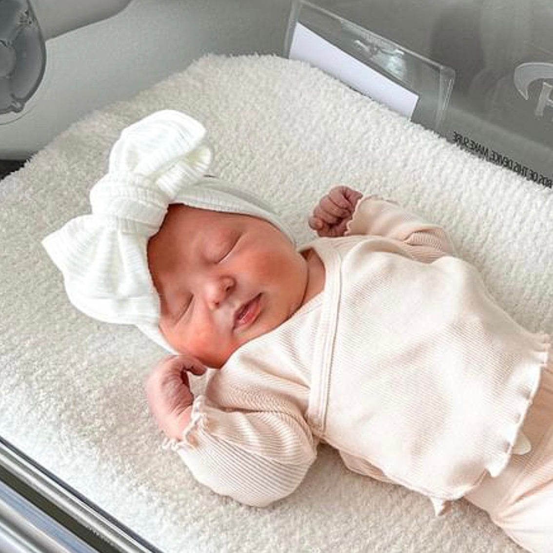 Newborn baby girl in a hospital cot, wearing a blush pink outfit and white ribbed oversized bow headband.