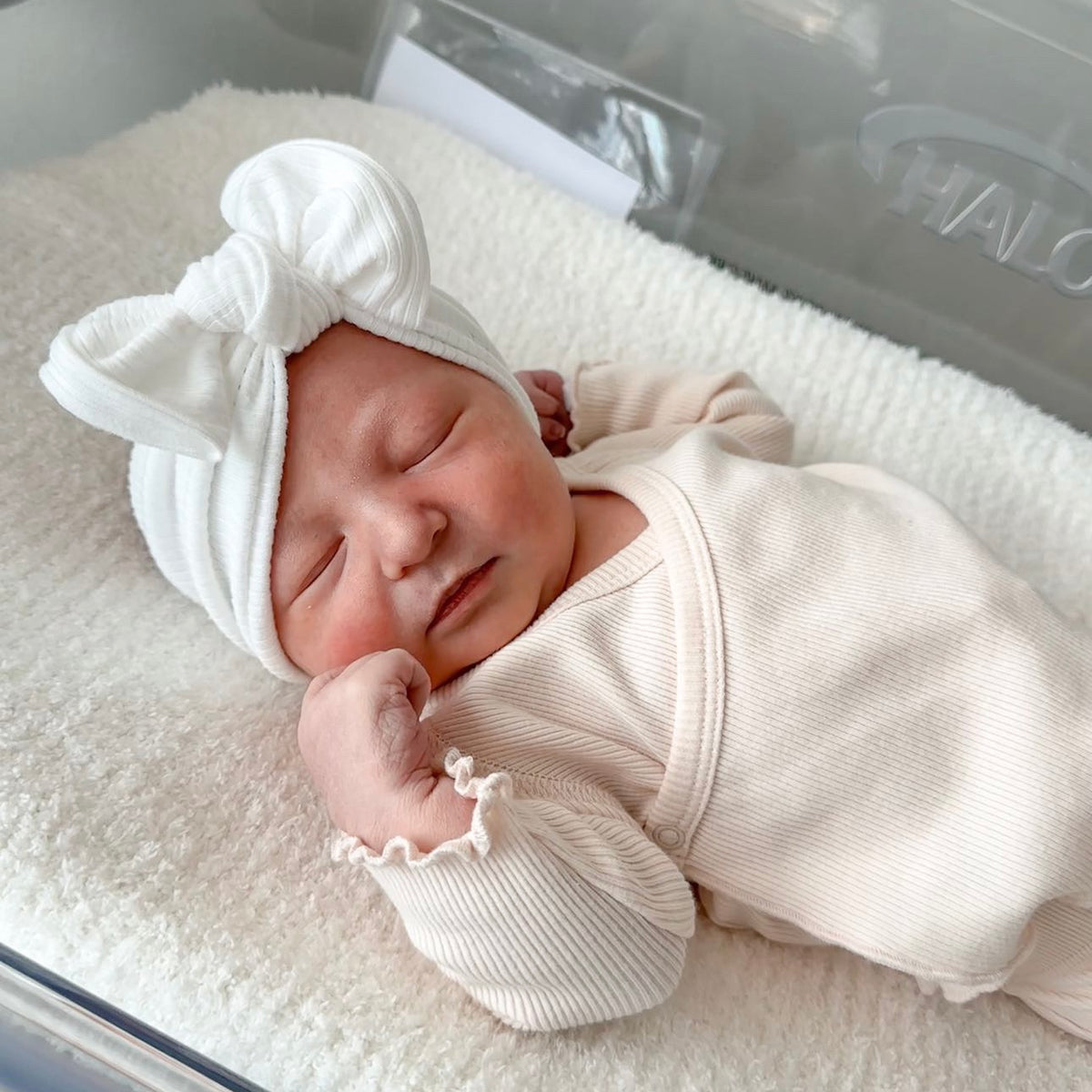 Newborn baby girl in a hospital cot, wearing a blush pink outfit and white ribbed top knot bow headband.