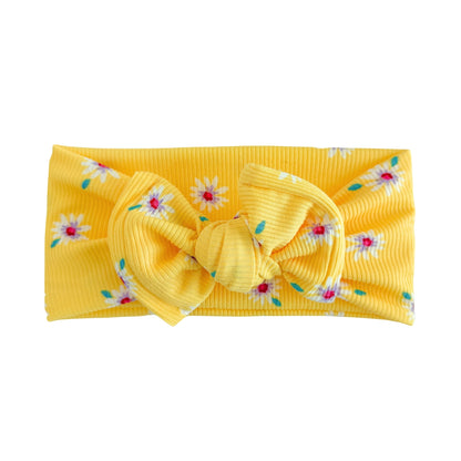 Oversized bow baby headband in bright yellow with white daisies print, crafted from ribbed knit fabric, displayed on a white background